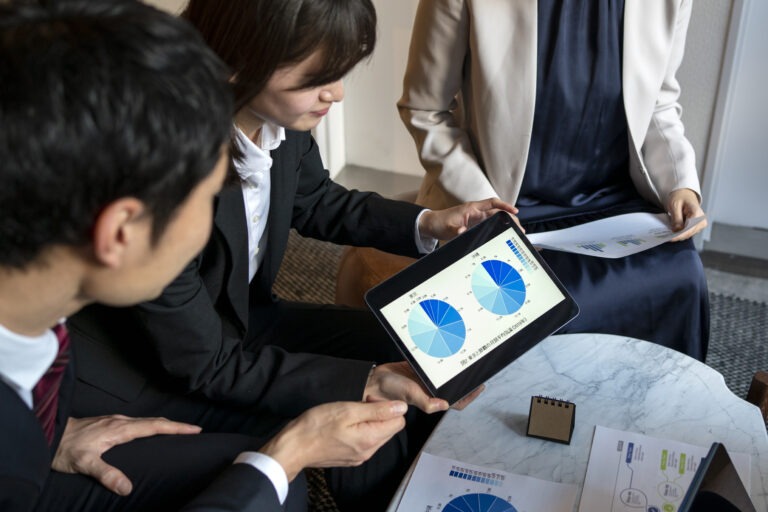Business professionals review data on a tablet during a meeting, focusing on pie charts and graphs, with documents and a notepad on a marble table.